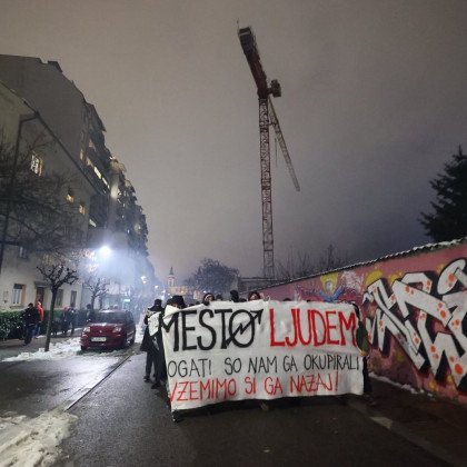 Večerna fotografija Trubarjeve ulice, viden je grafitiran zid tovarne rog na desni, skupina temno oblečenih oseb drži transparent z napisom MESTO LJUDEM / Bogati so nam ga okupirali / Vzemimo ga nazaj. V ozadju se vzpenja žerjav