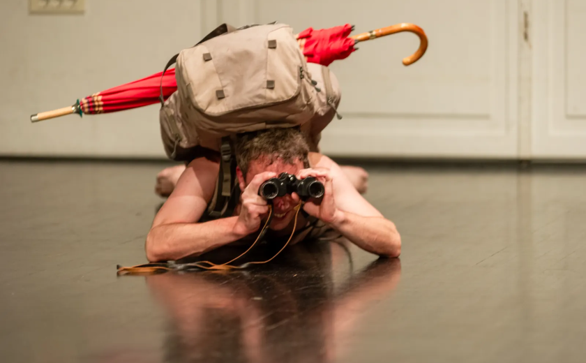 Performer Rok Kravanja na tleh z oprtanim nahrbtnikom, nekaj prav zadovoljno opazuje skozi daljnogled.